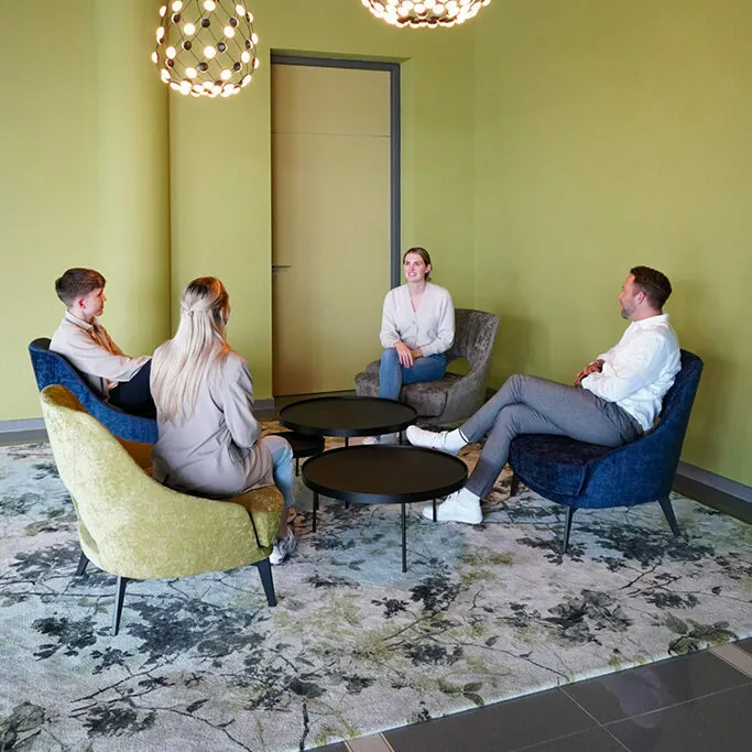 Four people sit on colorful chairs around a small round table in a modern room with green walls and decorative lighting.