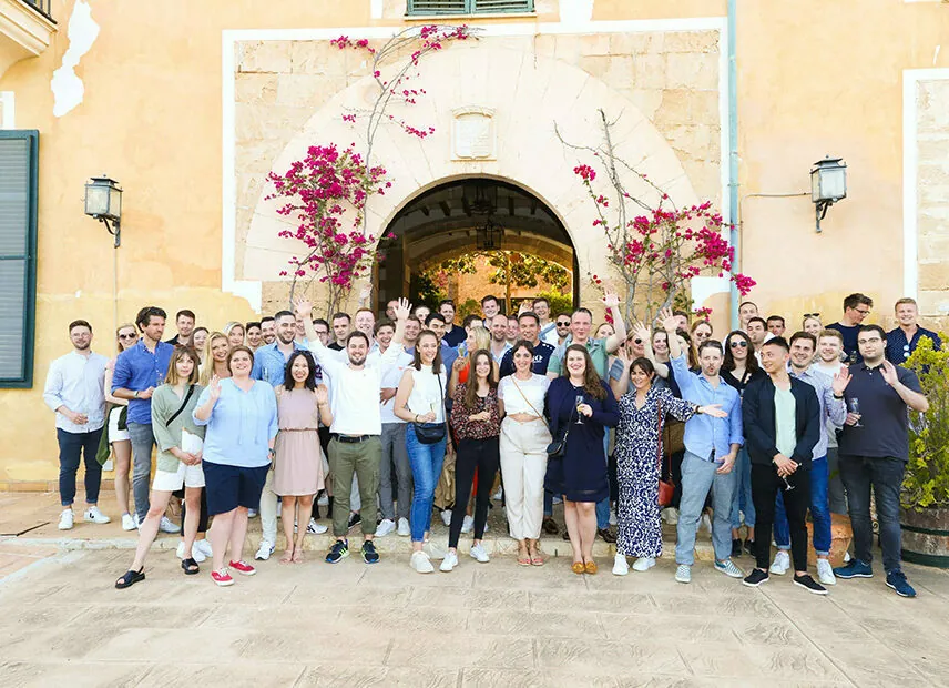 A large group of people posing for a photo in front of a mediterranean-style building with archway and bougainvillea. Many are smiling and holding drinks.