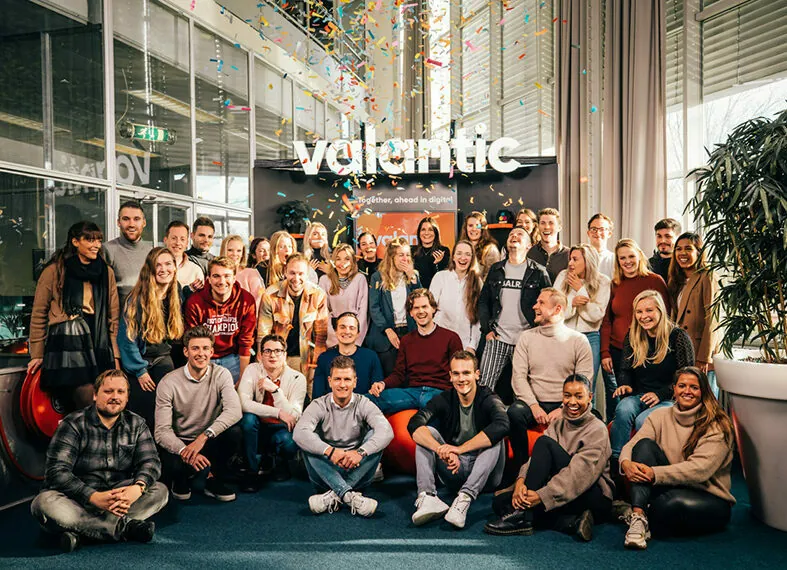 A group of people posing for a photo indoors in front of a "valantic" sign. There is colorful confetti falling from above. The group appears to be smiling and casually dressed.