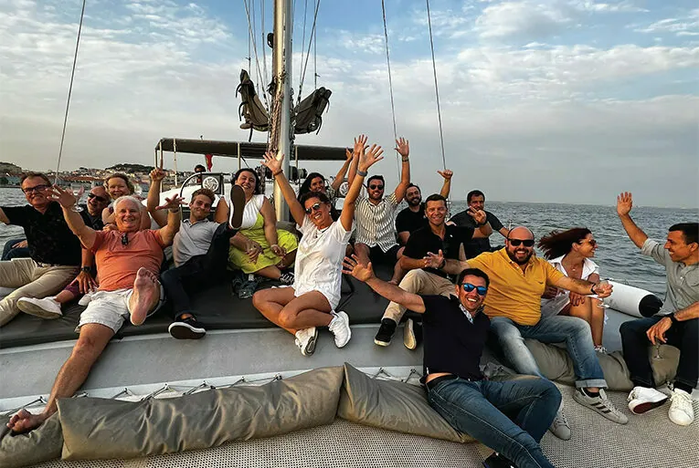 A group of people sitting on a boat deck, smiling and raising their arms. The sea and cloudy sky are visible in the background.