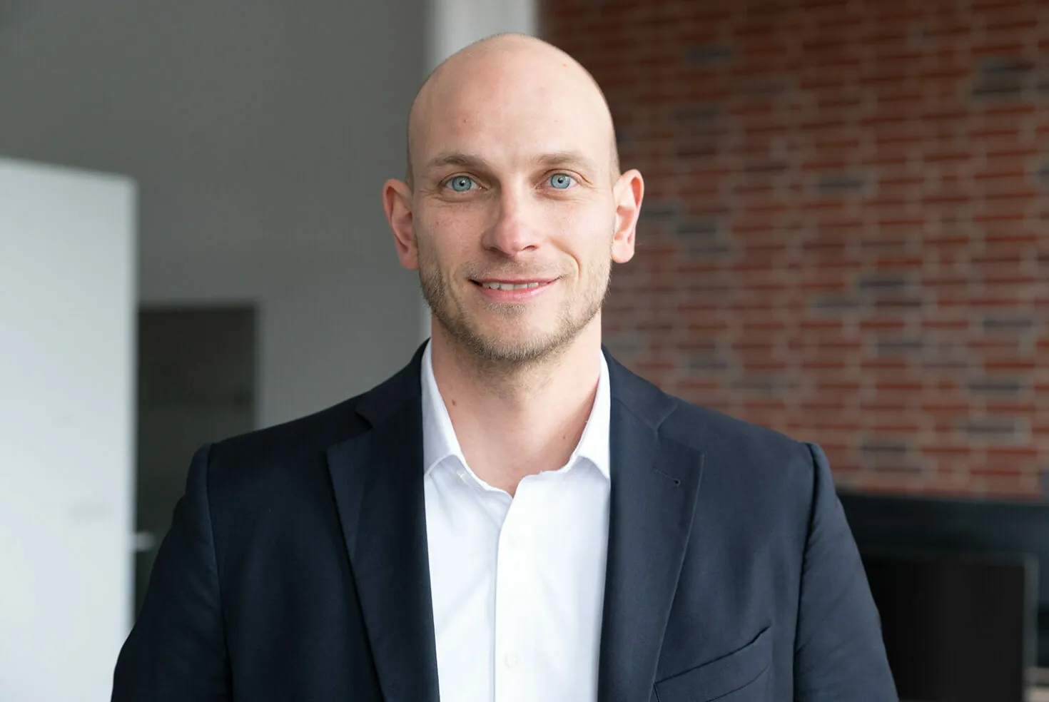 A person wearing a black suit jacket, white shirt, and smiling, stands indoors with a brick wall and a doorway in the background.
