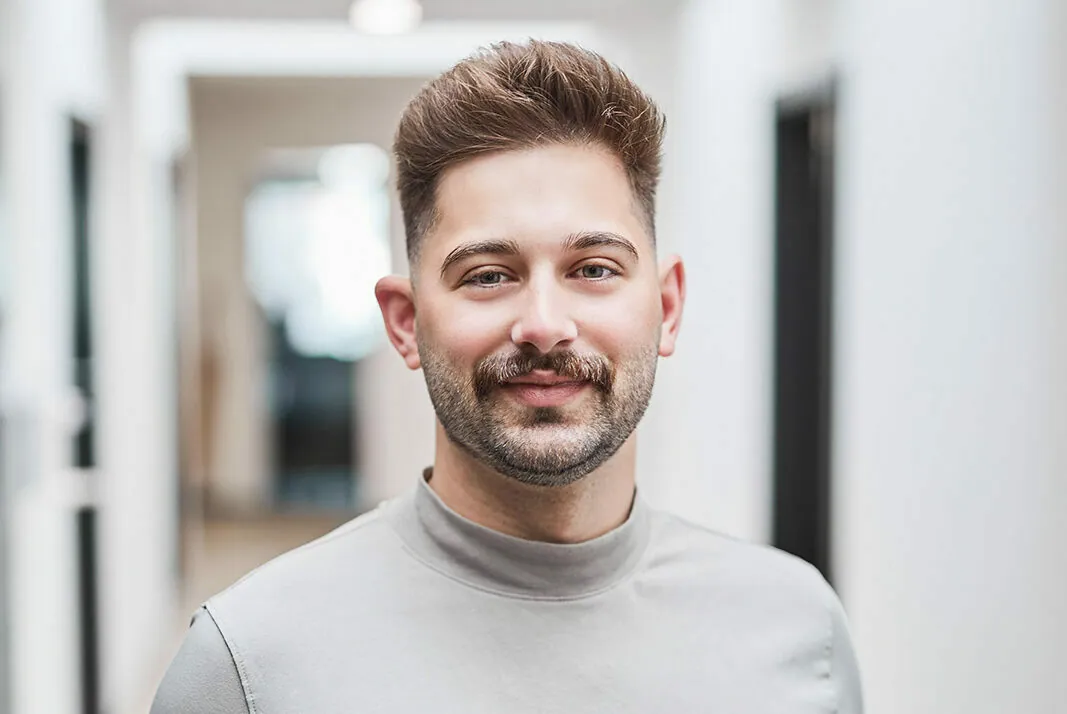 A person with short brown hair and a mustache, wearing a light gray shirt, is standing in a hallway with a neutral expression.