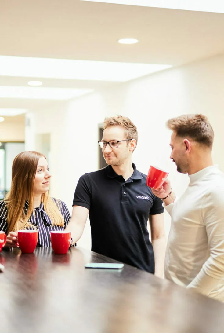 Three people are standing and talking around a table, each holding a red mug. One person is wearing a black polo shirt, and the other two are dressed in casual attire. There are more red mugs on the table.