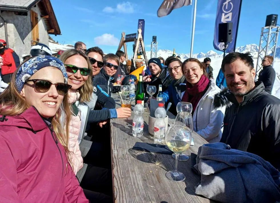 A group of people in winter clothing sits around a wooden table outside, with drinks on the table and snowy mountains in the background.