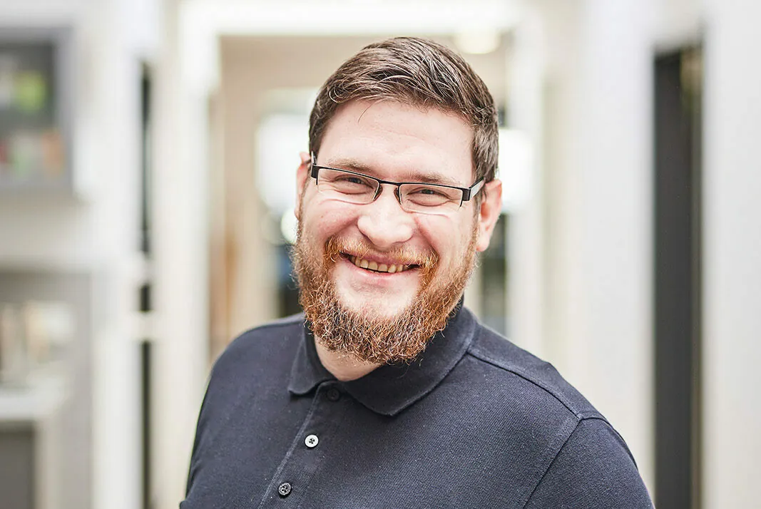 A man with a beard and glasses is smiling while standing in a bright hallway. He is wearing a dark polo shirt.
