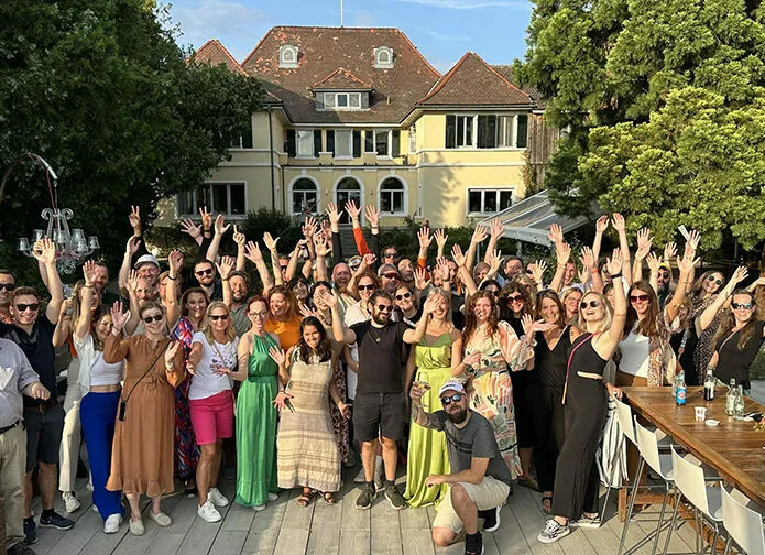 A large group of people in summer attire posing for a photo with their hands raised outside a building with trees and a clear sky in the background.