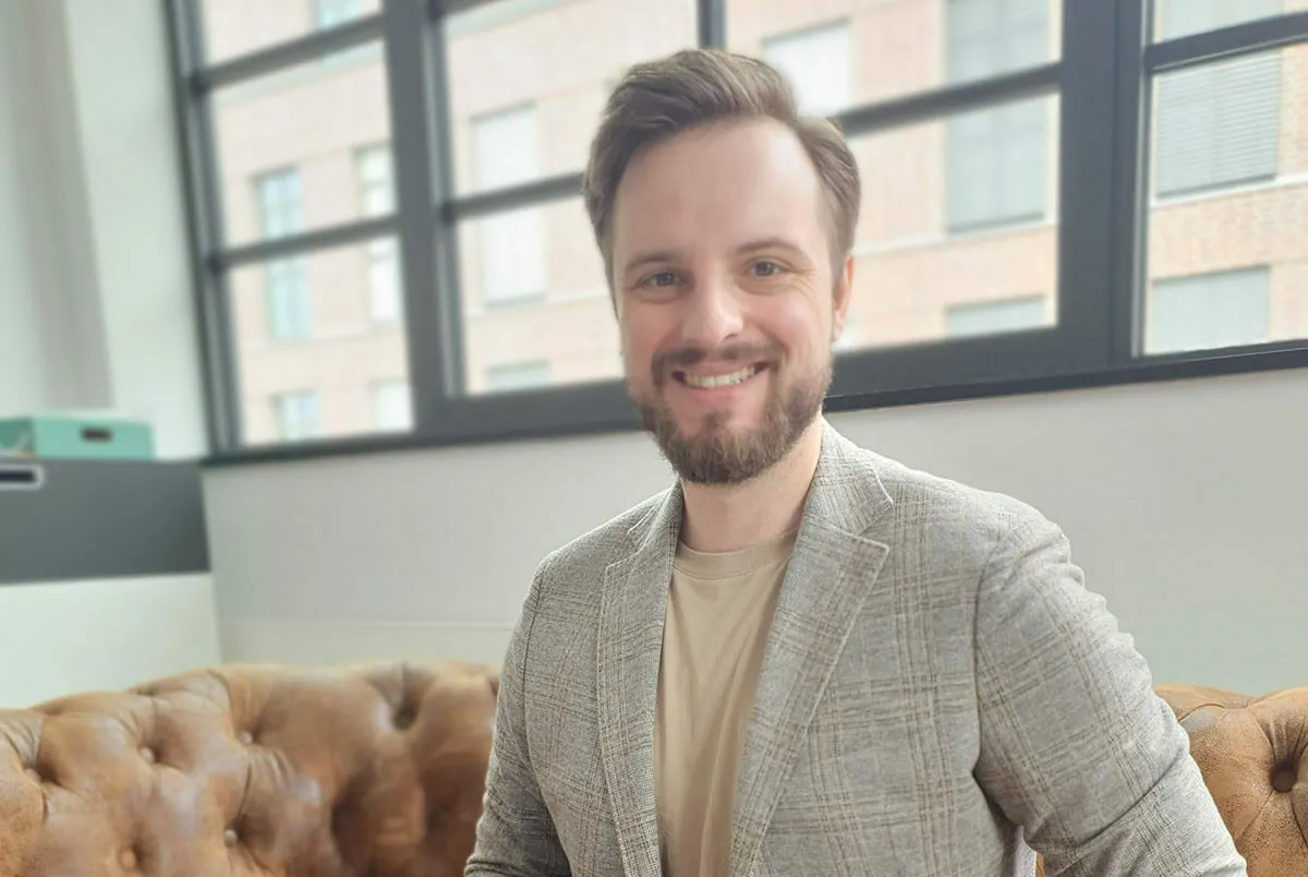 A man with a beard and short hair wearing a light gray blazer and beige shirt sits on a brown leather couch in front of large windows.