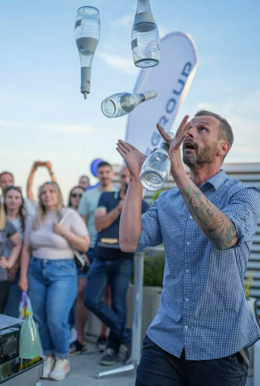 A man with tattoos and a checkered shirt juggles four glass bottles while an audience watches outdoors.