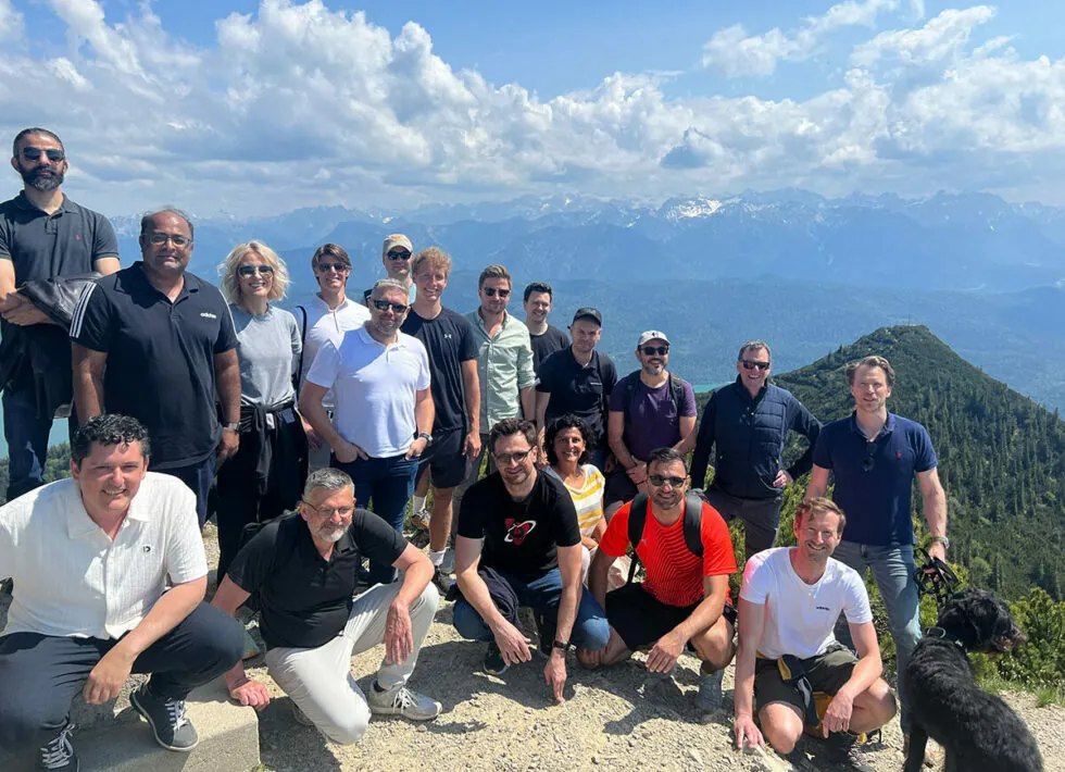 A group of 20 people posing outdoors on a mountain with a scenic backdrop of more mountains and a few clouds in the sky. A black dog is also present.