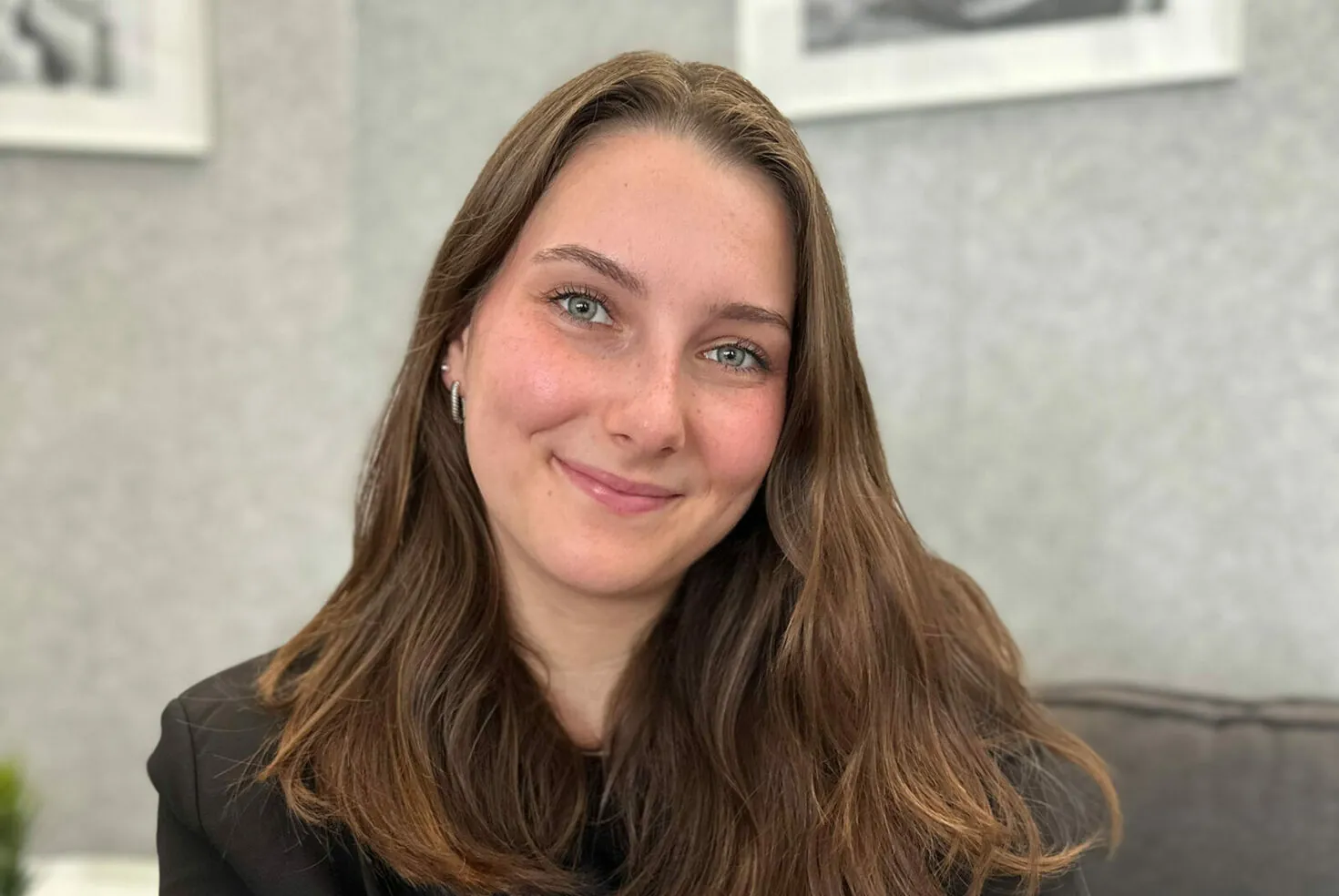 A woman with long brown hair and blue eyes smiles at the camera. She wears a black top and sits in a softly lit, neutral-toned room.
