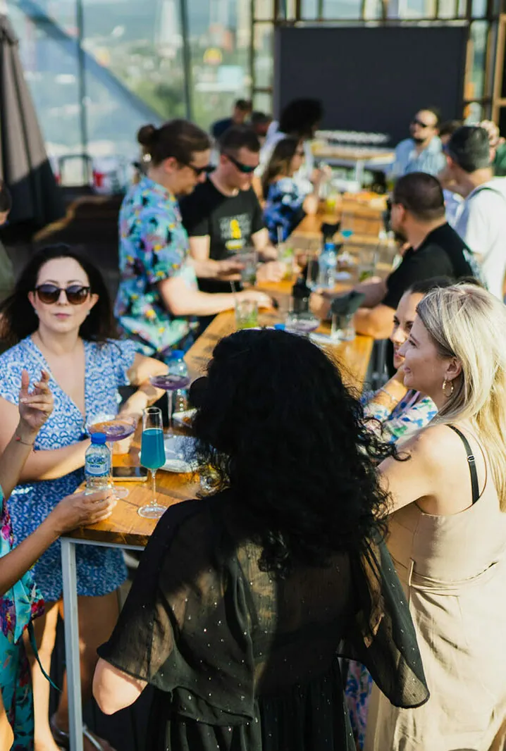 A group of people gather around a long wooden table outdoors, engaging in conversation and enjoying drinks.