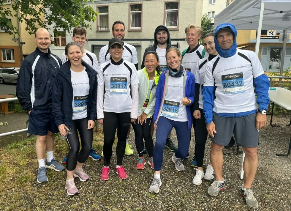 A group of people wearing athletic gear and numbered race bibs pose for a photo outdoors, with buildings and a tent visible in the background.