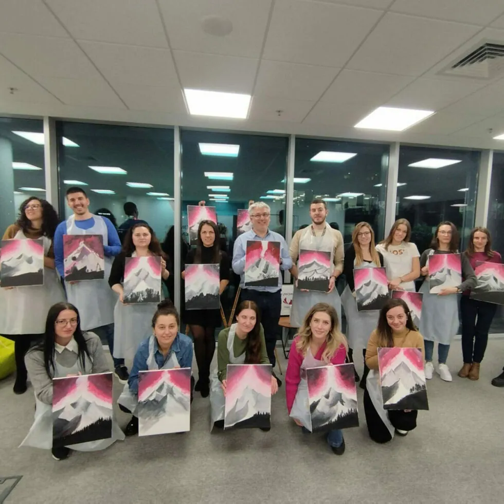 A group of individuals, each holding a painting featuring a mountain scene with pink and gray hues, pose together in an office setting with large windows in the background.