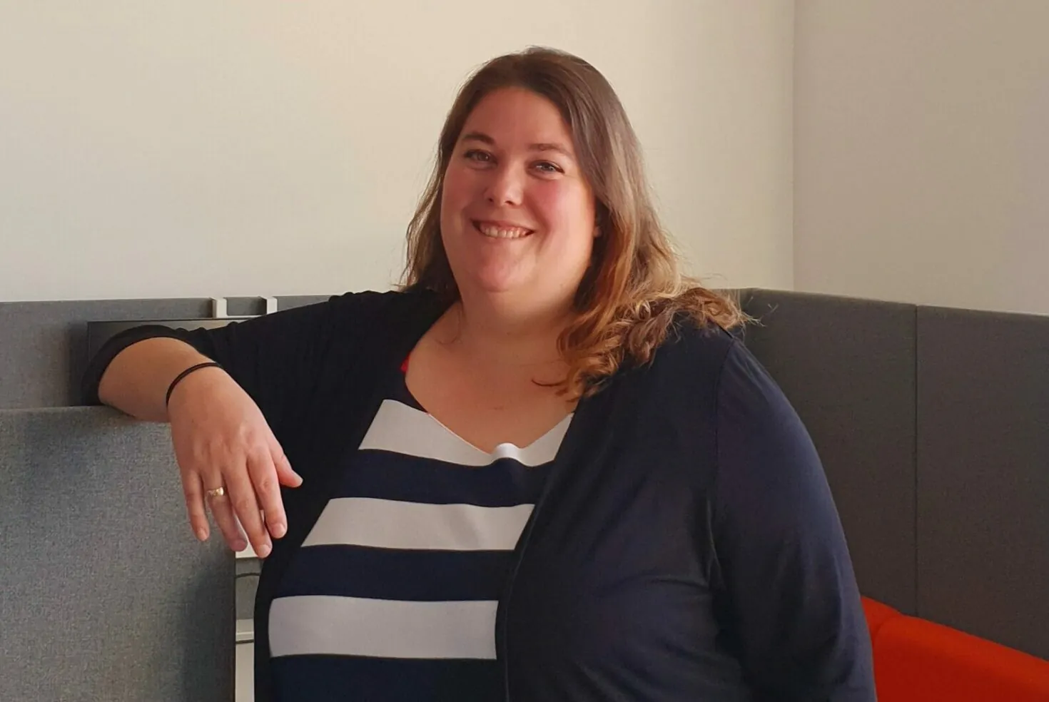 A woman with long, brown hair is smiling while leaning against a gray cubicle partition. She is wearing a black cardigan over a black and white striped top. The background is an office setting.