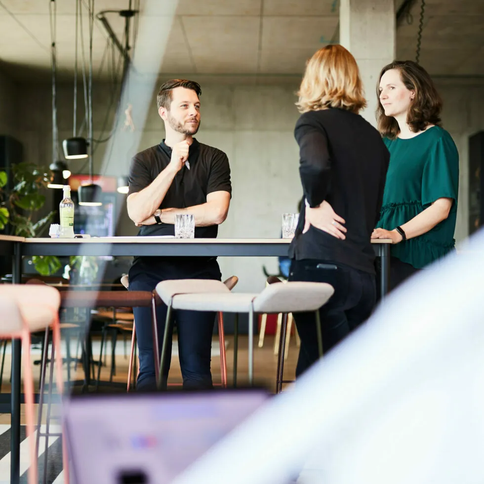 Three people are standing around a tall table in a modern indoor setting, engaged in conversation. One person has their hand on their chin, while the other two are facing them, listening attentively.