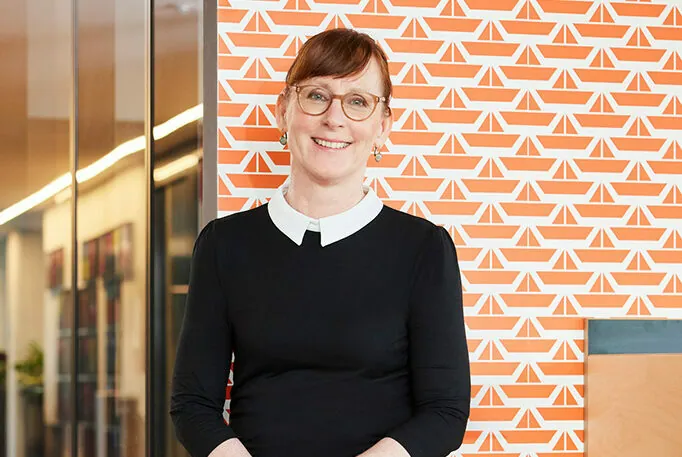 A person with glasses and a black dress with a white collar standing in front of an orange wall with white geometric patterns.