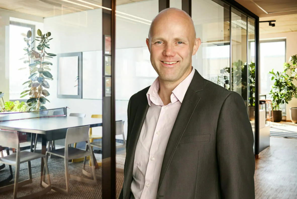 A bald man in a business suit smiles while standing in front of a glass-walled office room filled with tables, chairs, and plants.