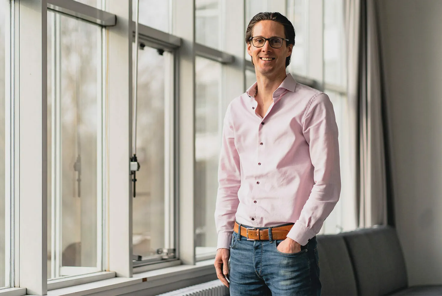 A man wearing glasses, a light pink button-up shirt, and blue jeans stands near a bright window, smiling.