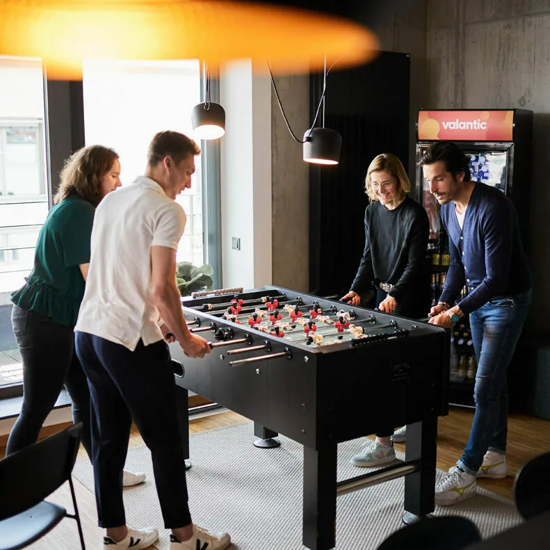 Four people are playing foosball together in a room with modern lighting. A window in the background lets in natural light, and there is a vending machine with the brand name "valantic" on it.