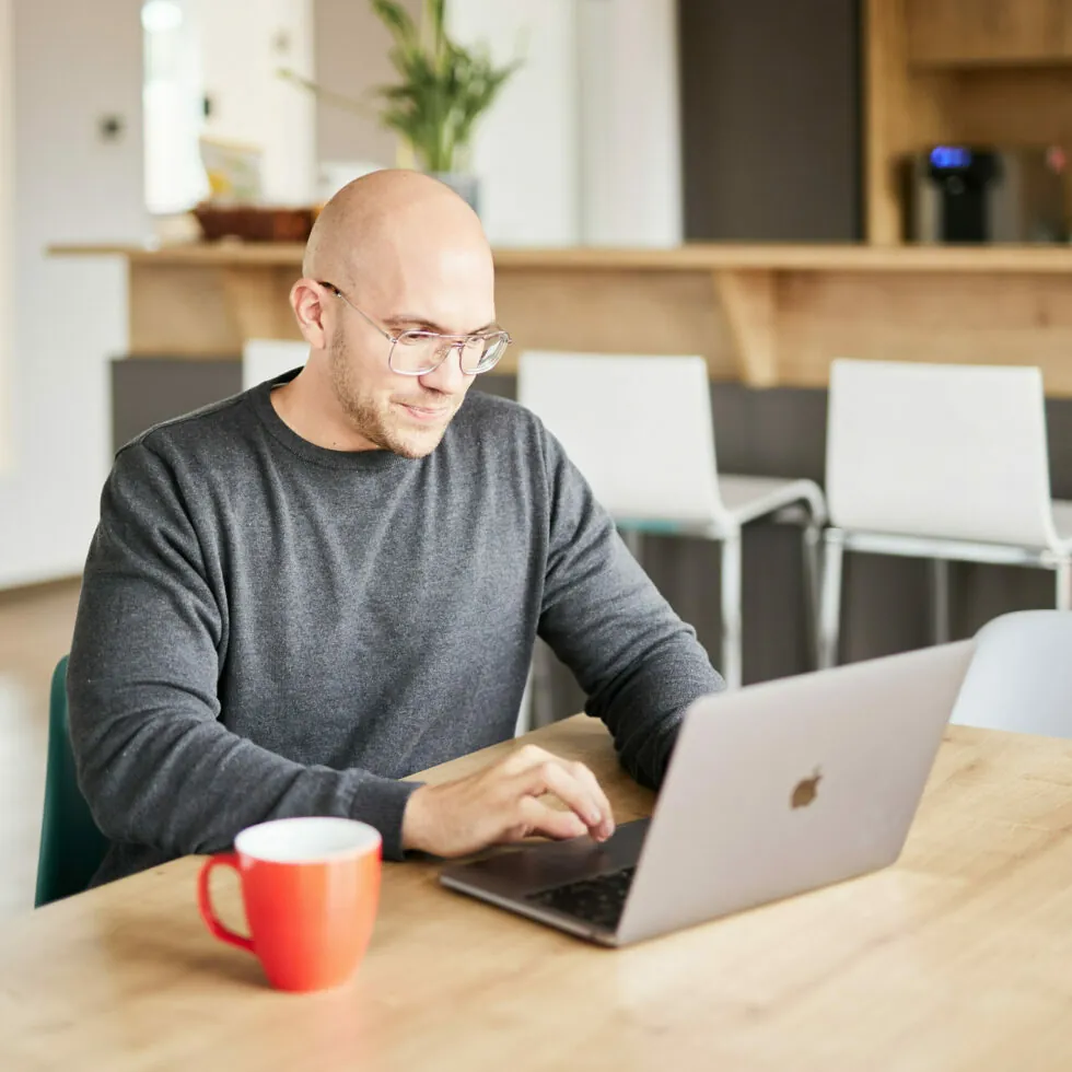A person with glasses using a laptop at a wooden table with a red mug nearby. An open space with chairs and a counter is in the background.