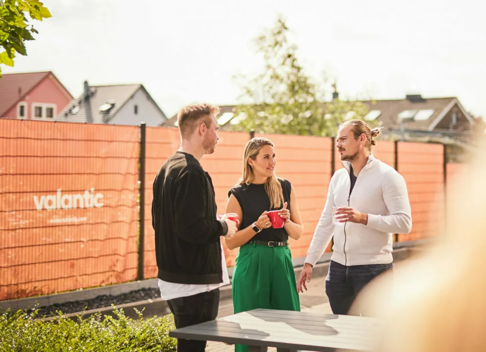 Three people stand outdoors, having a conversation near an orange fence with the word "valantic" on it. The person in the middle holds a red cup. Houses and trees are visible in the background.