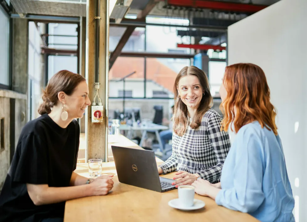 Three women are sitting at a wooden table in a modern office, engaged in a conversation. A laptop is open on the table, and a coffee cup is placed in front of one of the women.