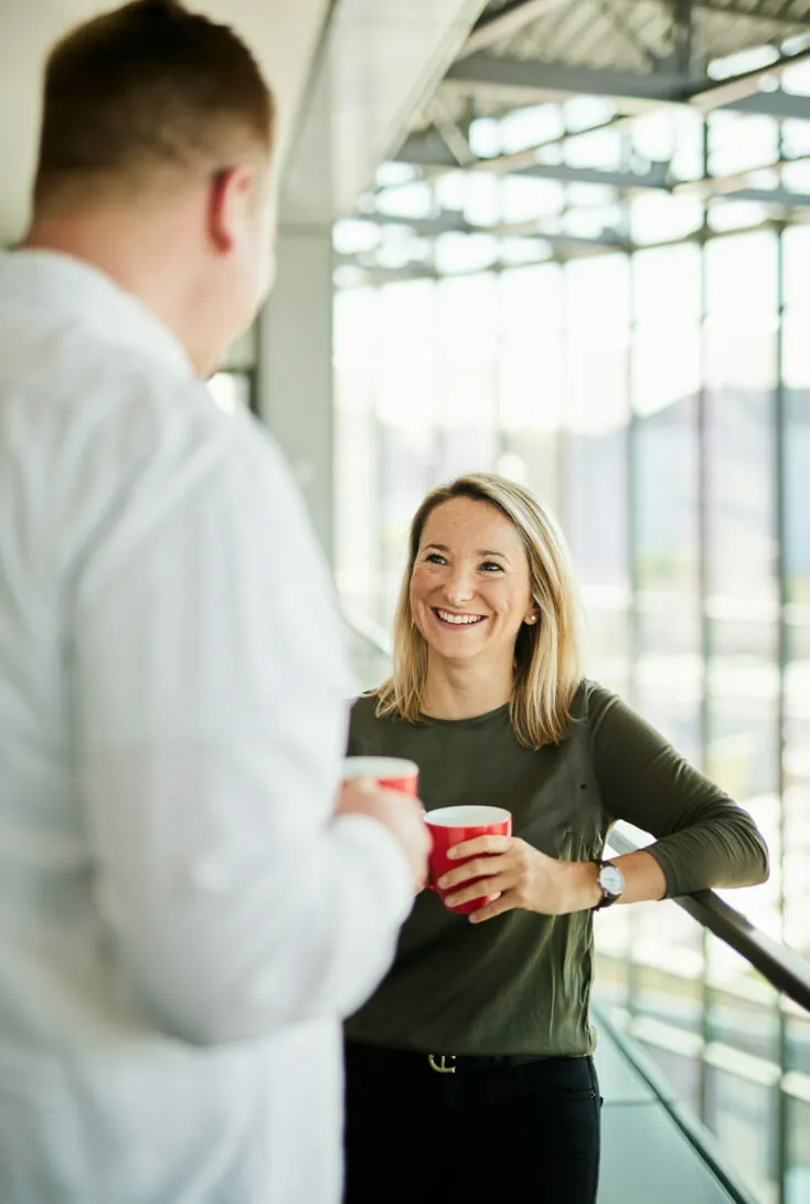Two people are standing indoors, holding red mugs and talking. One person is wearing a white shirt and the other a green top. They are near large windows with a blurred outdoor view in the background.