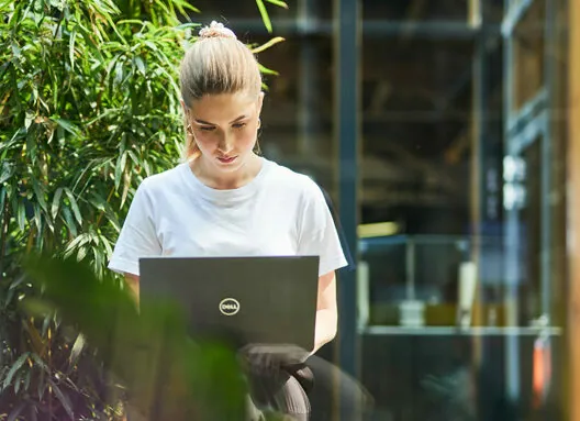A woman in a white t-shirt works on a Dell laptop while standing near plants indoors.