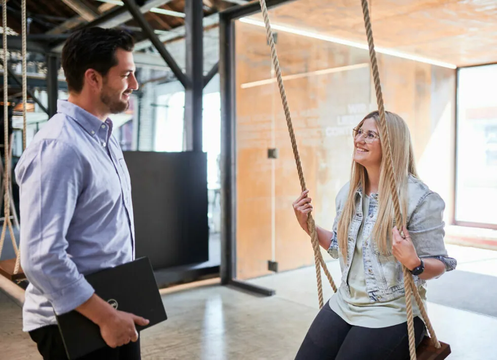 A man standing and holding a laptop talks to a woman sitting on a swing inside an office space.