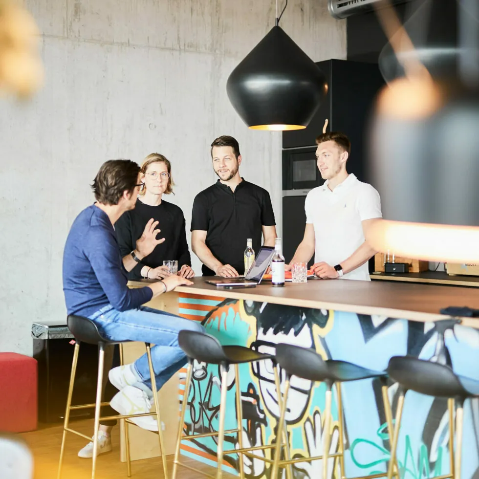 Four people engaging in conversation at a modern bar counter with a colorful graffiti design. Three are standing behind the bar, while one person is seated on a barstool in front.