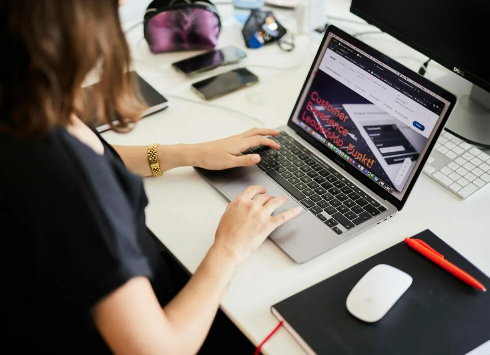 A person working on a laptop at a desk, with a webpage open on the screen. A smartphone, mouse, notebook, and other items are also on the desk.