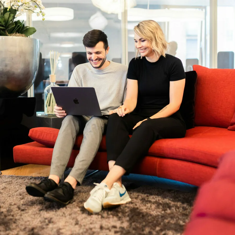 Two people, a man and a woman, sit together on a red couch, sharing a laptop. They appear to be engaged and smiling.