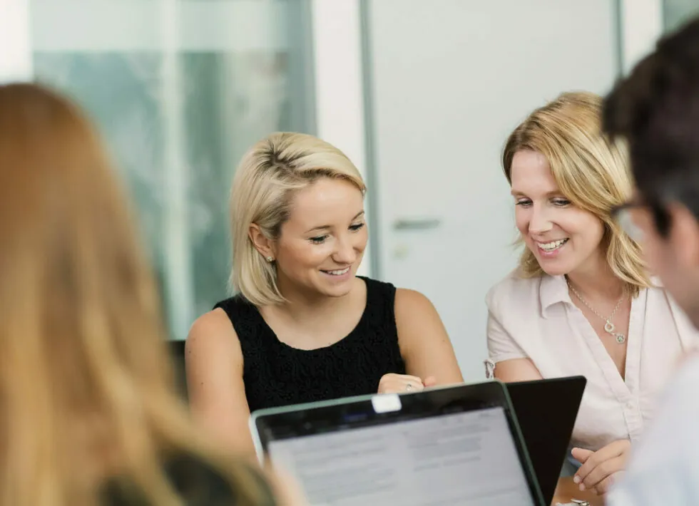 Two women are sitting at a table, engaged in conversation and looking at a laptop. There are two other people partially visible in the foreground, also focused on their laptops.