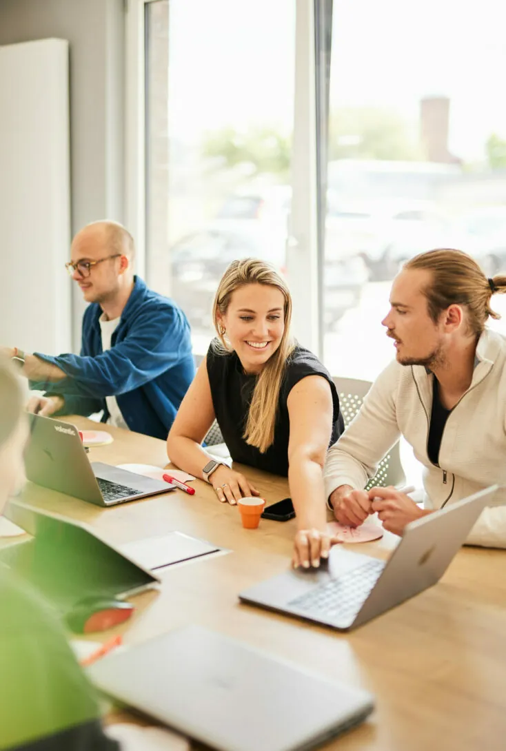 A group of three people are sitting at a wooden table, engaging with laptops and notepads in a bright room with large windows.