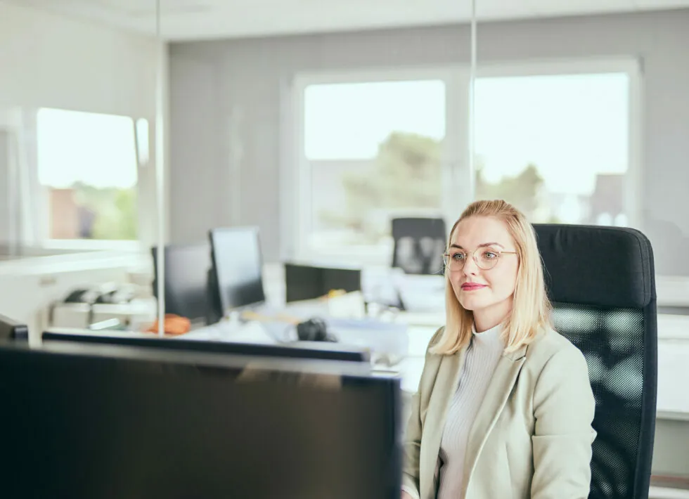 A woman with glasses sits in an office chair, looking at a computer monitor. The office space around her is bright with multiple desks and computer screens.