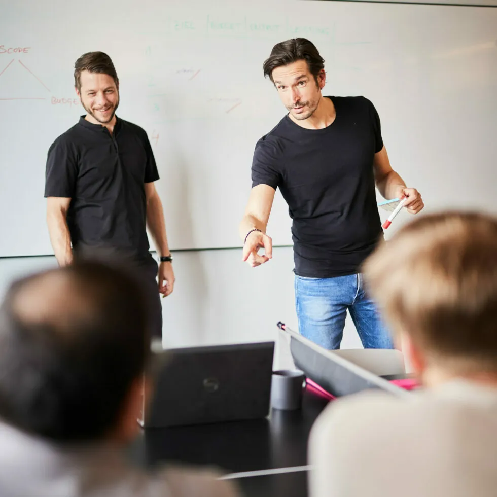 Two men lead a business meeting in a modern office. One man points at a laptop screen while the other stands nearby, both facing two seated individuals. A whiteboard with notes is in the background.