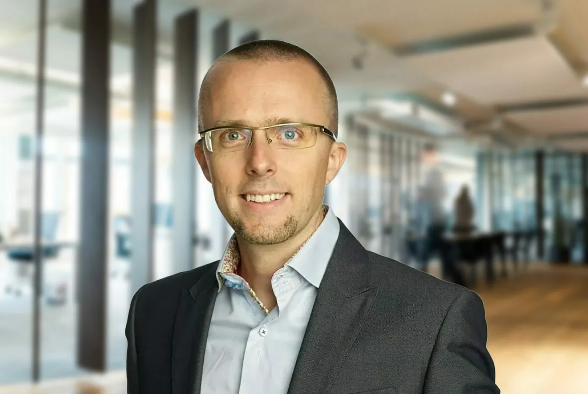 A person with glasses, wearing a light blue shirt and a dark blazer, stands in an office space with desks and glass partitions.