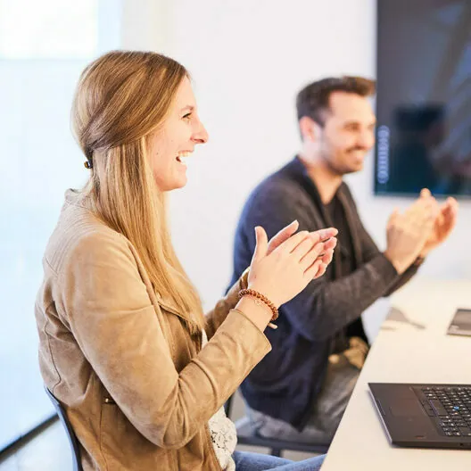 Two people are sitting at a table, clapping and smiling. A laptop is open in front of them. The background is a light, indoor setting.