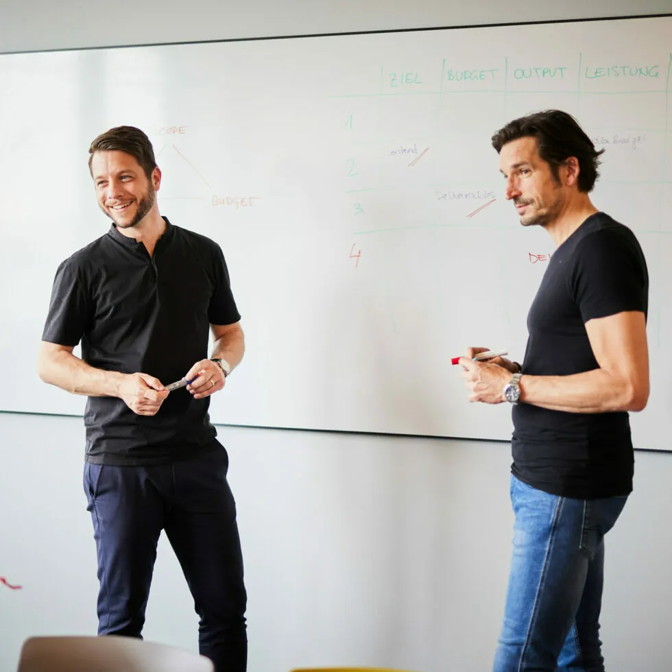 Two men standing and discussing in front of a whiteboard filled with text and charts. One man is holding a marker, and they both appear to be engaged in a conversation.