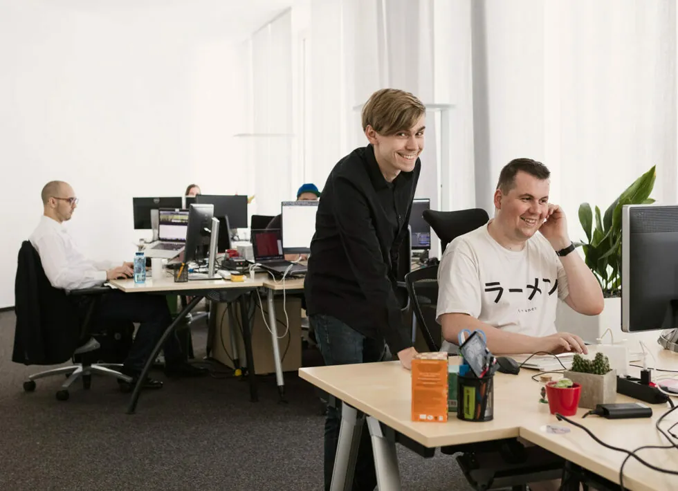 Three people are working in an office with two men interacting at the front desk and another man working at a computer in the background.