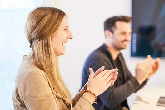 Two people, a woman in the foreground and a man in the background, are seated and clapping. Both appear to be engaging in a positive discussion or meeting.