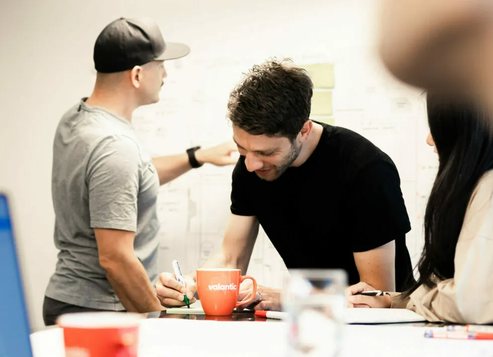 Three people collaborate in a meeting room. One person in a cap points at a whiteboard, while two others take notes. A red mug and scattered papers are on the table.