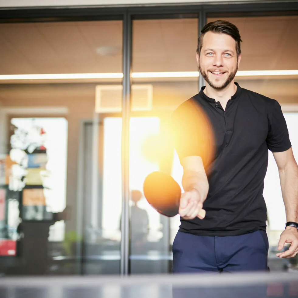 A man in a black shirt plays table tennis, mid-swing, with a smile on his face in an indoor setting with glass doors in the background.