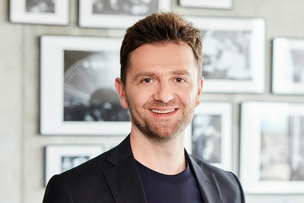 A person with short, dark hair and a beard is smiling, standing in front of a wall adorned with framed black-and-white photographs. They are wearing a black jacket over a dark shirt.