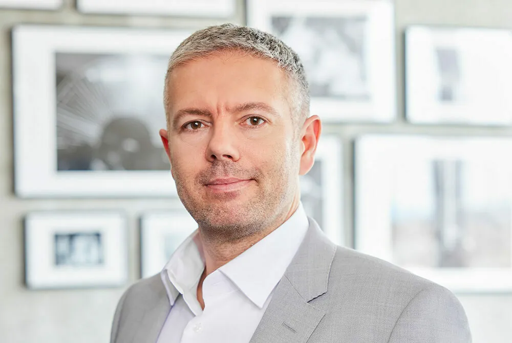 A man with short gray hair wearing a light gray suit jacket and white shirt stands in front of a wall with several framed black and white photos.