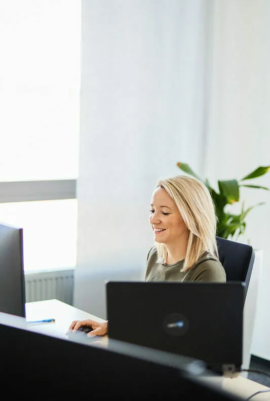 A person with long blonde hair is sitting at a desk, working on a laptop, in a bright office space with a plant in the background.