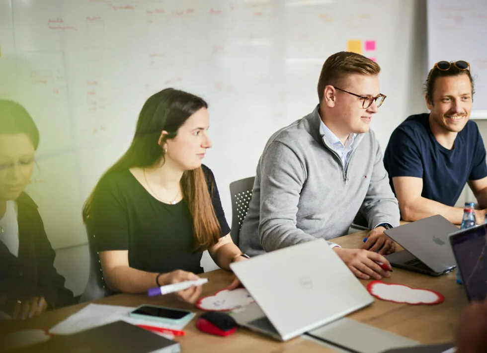 Four people are sitting at a table with laptop computers, engaged in discussion in a workspace with a whiteboard in the background.