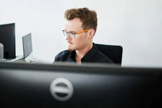 A man wearing glasses is seated at a desk and working on a computer. Multiple screens are visible in the scene.