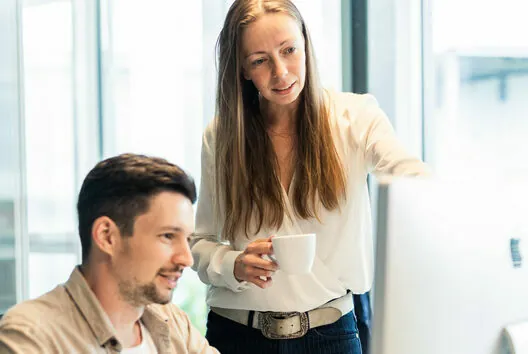 A woman holding a mug points at a computer screen while a man sits and looks at the screen. They appear to be working together in an office setting.