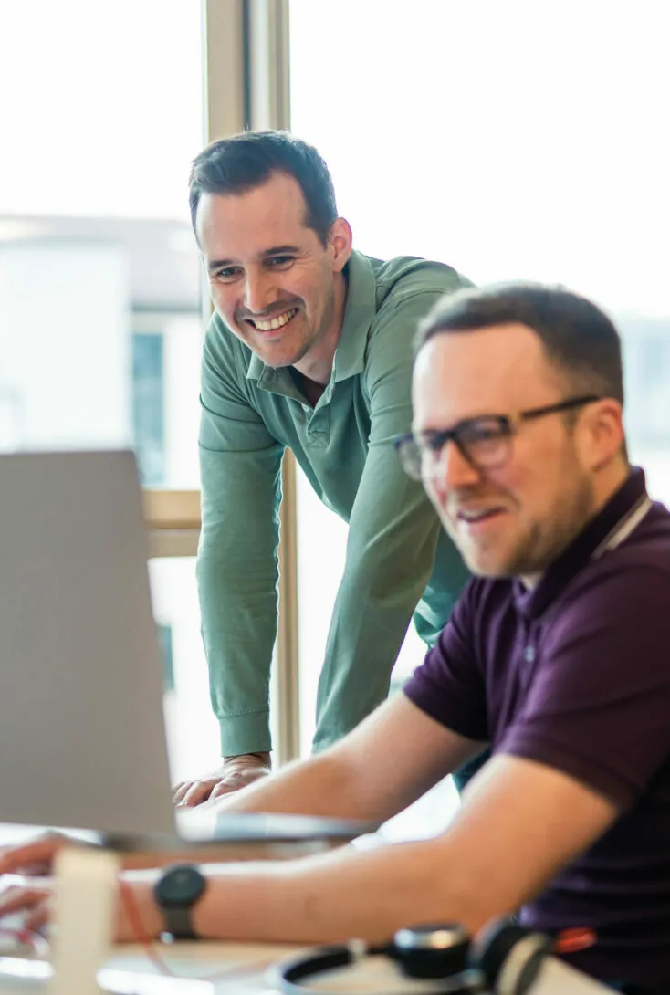 Two men working together at a computer, with one standing and leaning over the desk and the other seated, both smiling.